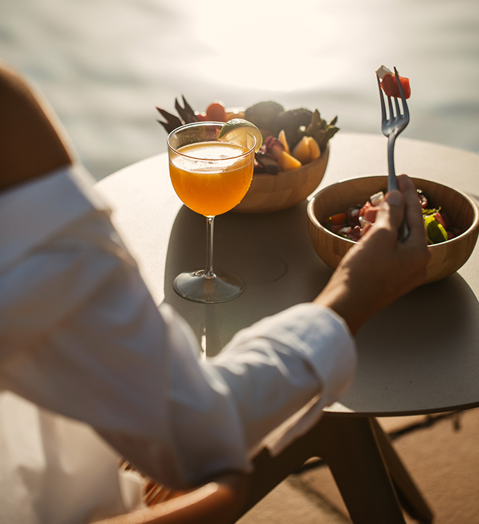 Orange cocktail on a small round table with a bowl of fruit and a salad with a woman's hand holding a fork with salad on it.