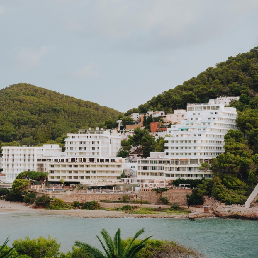 Large white hotel building on a sandy coastal beach with green mountains in the background