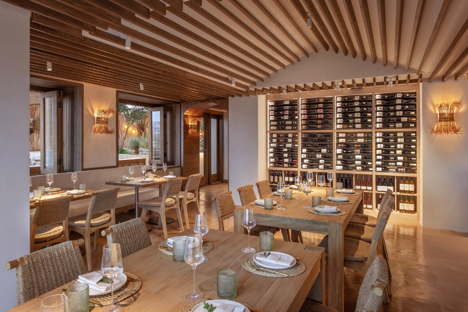 Interior of Casa Madera restaurant with long wooden ables and wicker chairs, and a wall with wine bottles on it