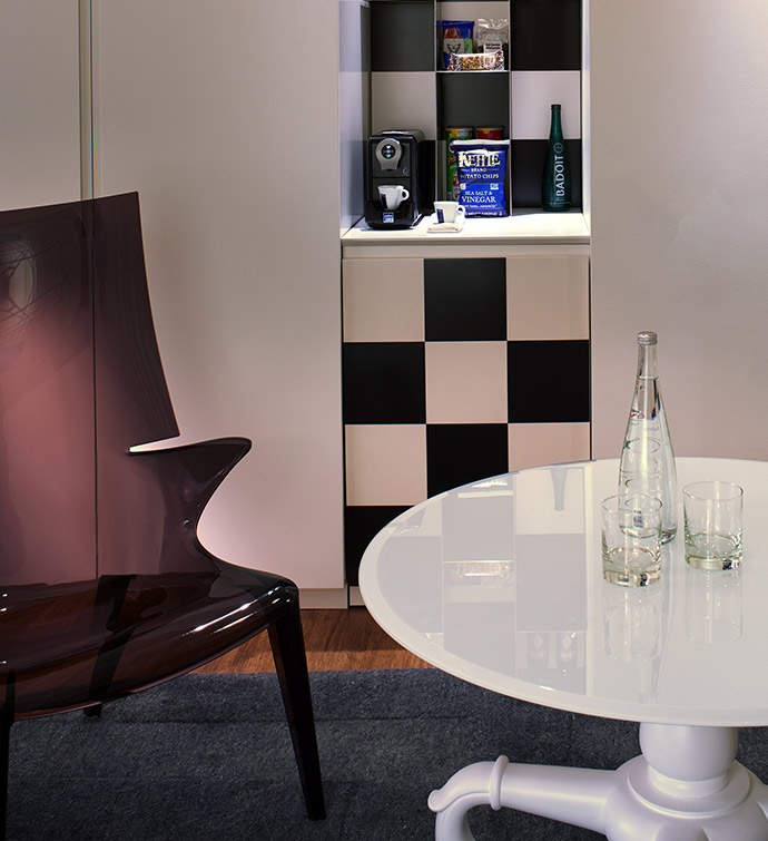 A corner with a coffee maker, table with water on it, and a clear chair.