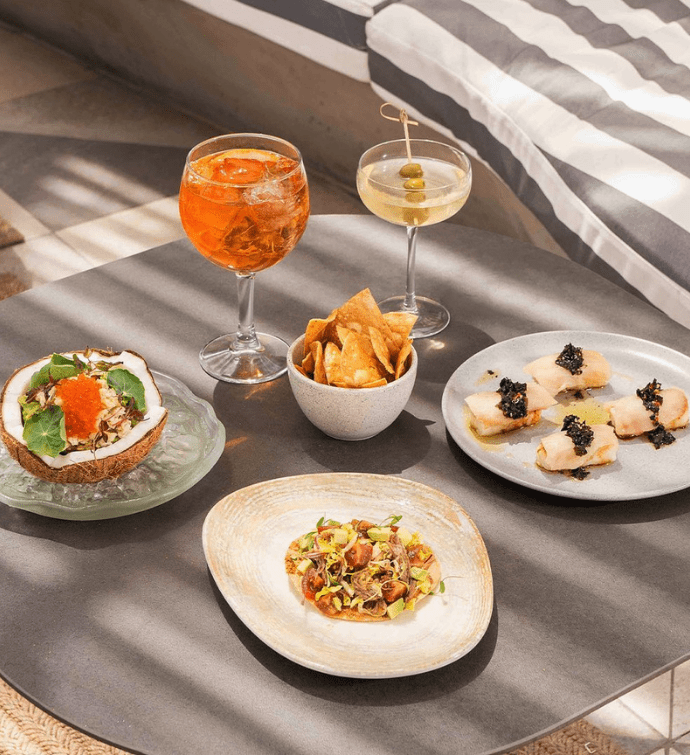 Three plates of appetizers on a grey placemat with a bowl of chips and two cocktails in the background