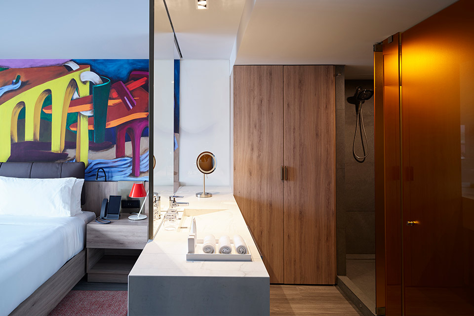 Hotel bed with a colorful wall mural, and bathroom with sink.