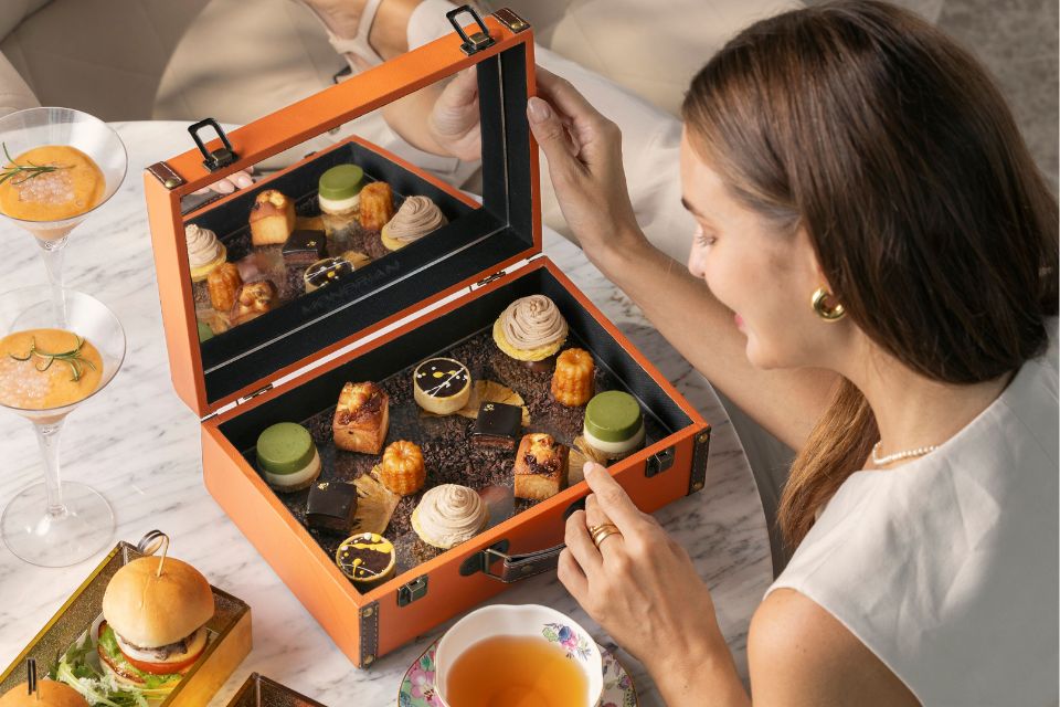 An intricate tea box filled with tea pastries is opened by a woman sitting at a white table with cocktails and cup of tea in the background