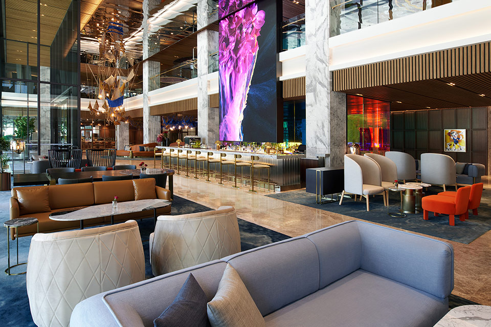 Large lobby area with grey sofa in the foreground and various seating in the background with an elaborate bar and elaborate art work hanging throughout.