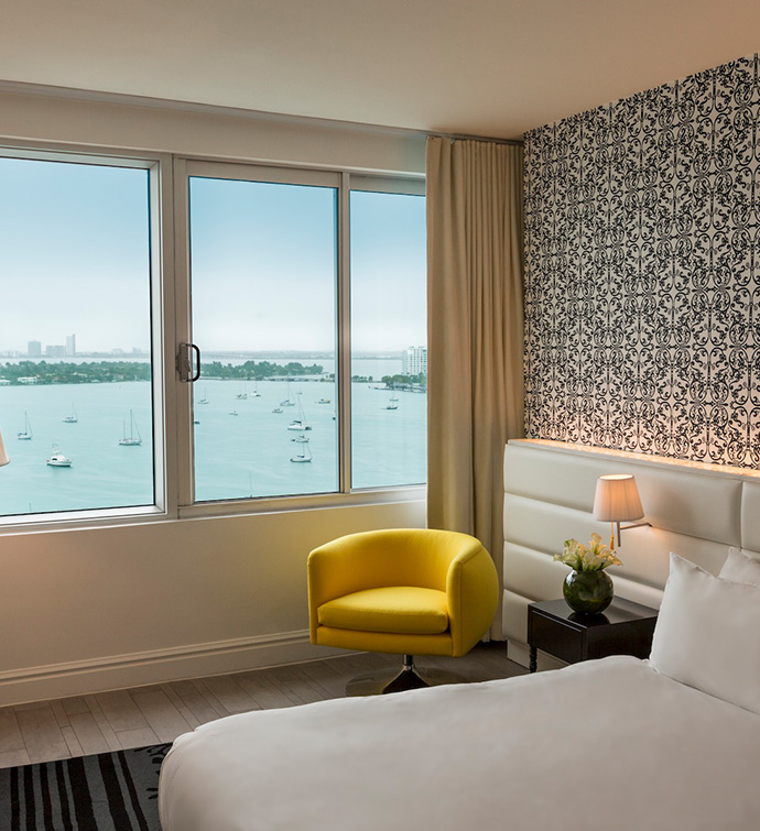 Hotel room with bed, yellow chair, and window overlooking the water.