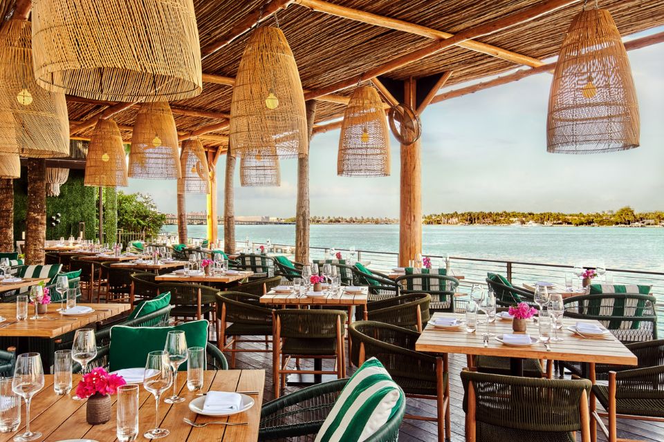 Large outdoor deck overlooking the beach with light wooden square tables, outdoor chairs with green and white striped pillows, and rattan hanging lights