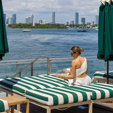 Woman in white outfit pours a glass of rose sitting on a green striped lounge chair looking out to the ocean