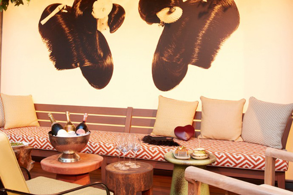 Outdoor upholstered banquet with pillows alongside several mismatched accent tables, one with a silver bowl holding three bottles of champagne