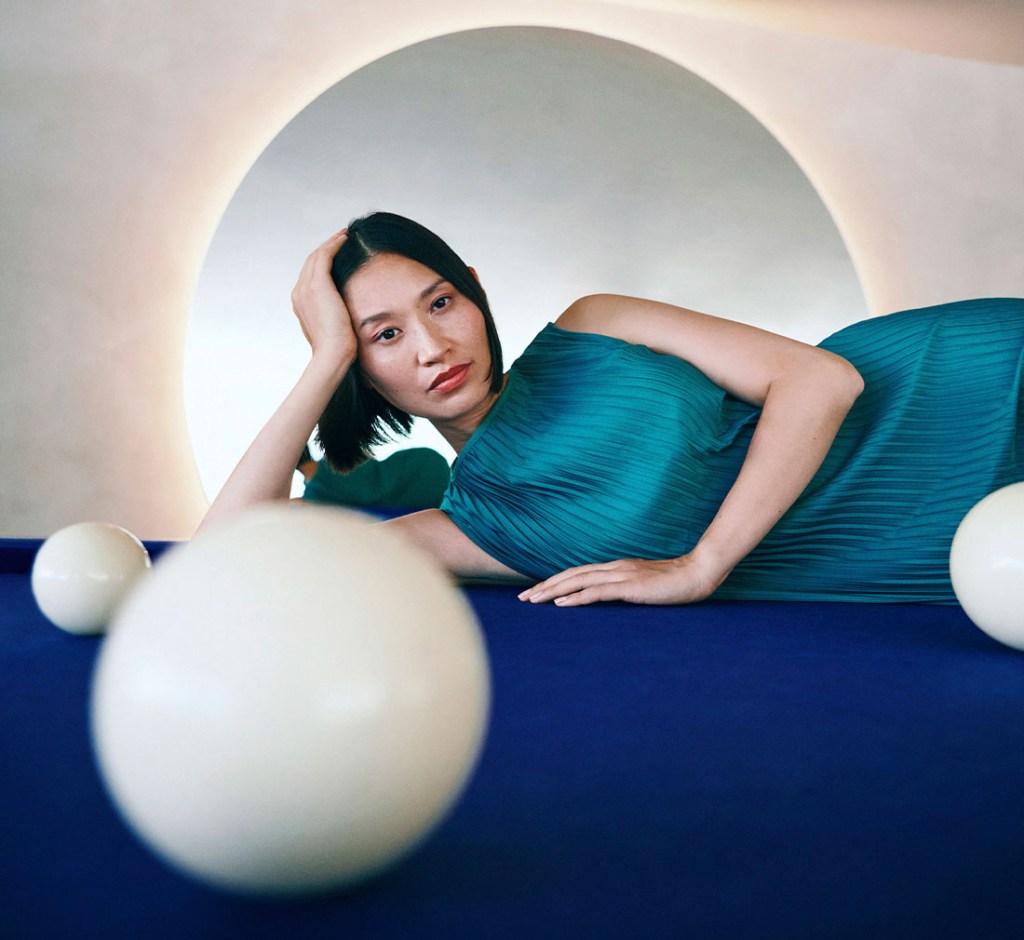 A woman in a teal dress leaning on a blue pool table with white pool balls