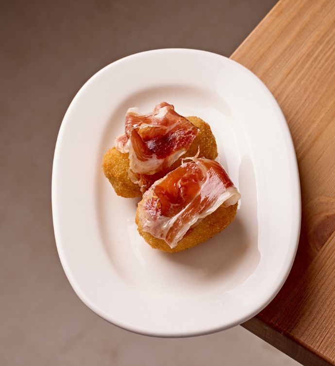 Two croquetas topped with meat on a white oval plate on a light wooden table top