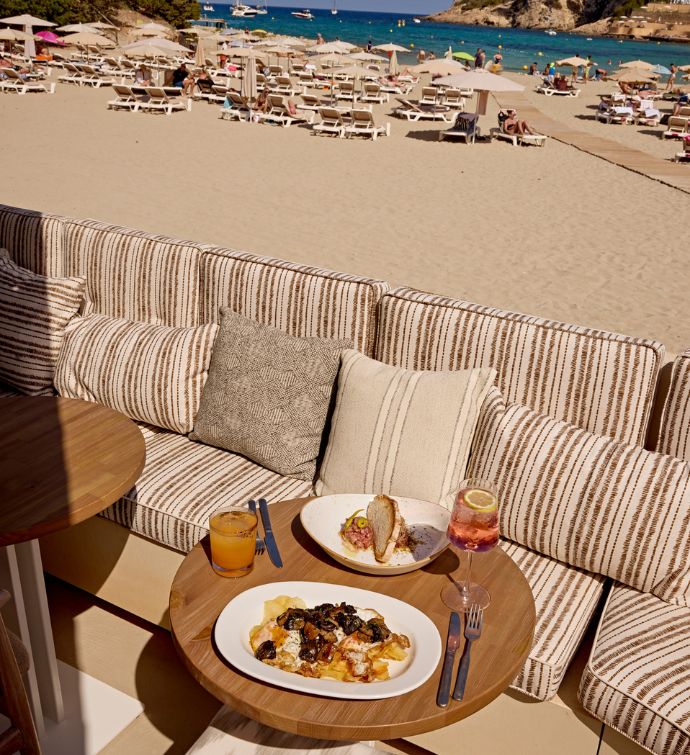 Small round table with cushion striped seating and dishes and drinks on it with a beach in the background