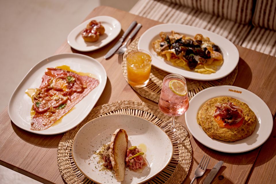 Five dishes on various sized and shaped plates sit on a wooden table with two cocktails garnished with fruit slices