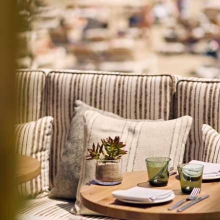 Close up image of a small wooden round table with plant, cups, and plates on top and a cushioned booth with extra striped pillows behind it and a beach blurred in the background