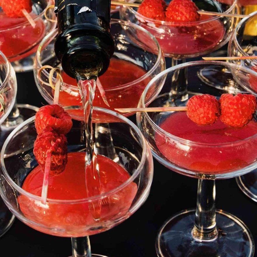 champagne being poured into a coup glass garnished with raspberries, surrounded by 4 other full coup glasses