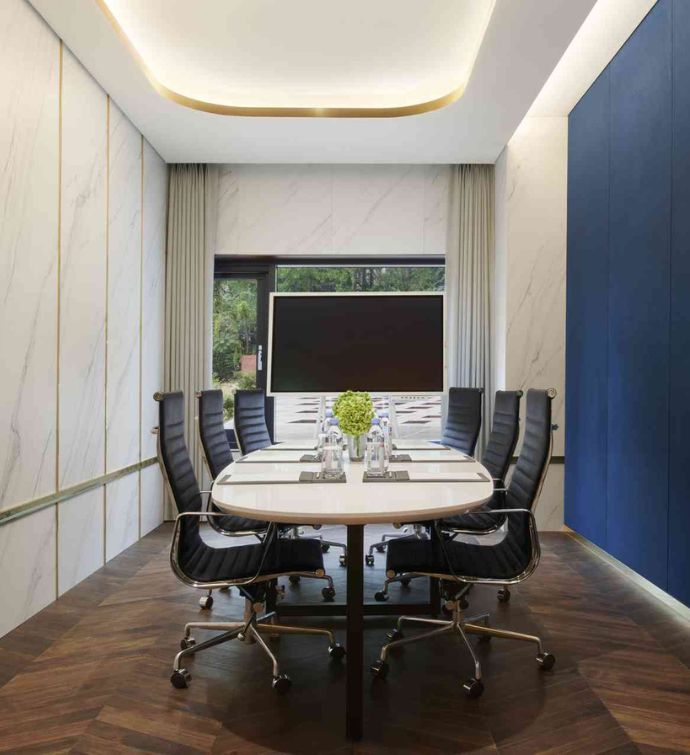 Small conference room with a light wooden oval table and rolling desk chairs on either side with a large screen at the front of the room and one blue wall