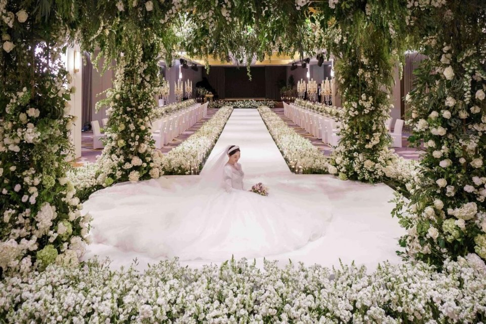 Woman in a large white wedding dress sits amongst lots of flowers in a wedding venue