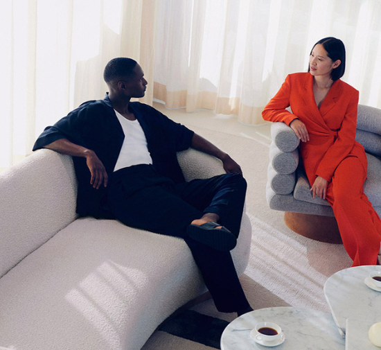 A man dressed in a black two piece outfit, and a woman in an orange jumpsuit, sitting in chairs enjoying a cup of coffee.