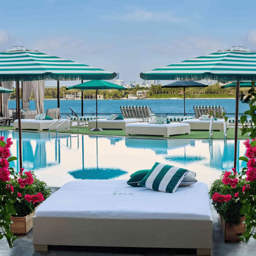 Daybed by pool with green and white striped pillows, surrounded by pink flowers, with Biscayne Bay in the background