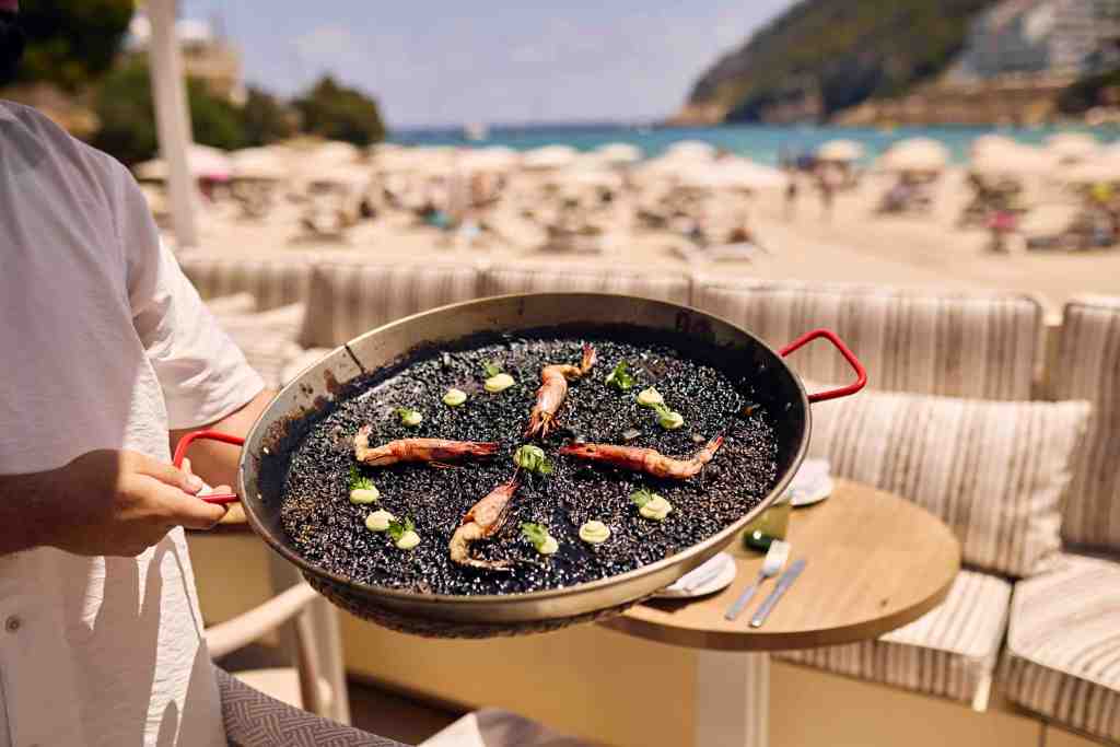 Large pan of seafood paella held up by a chef's arms with a beachside restaurant in the background