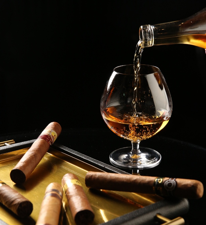 Pouding liquor into a brandy glass near a tray of cigars 