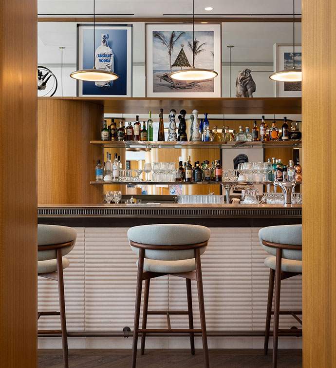 Bar with grey and wooden frame stools, shelves of liquor and glassware, pendant lights, and two wooden columns in the foreground