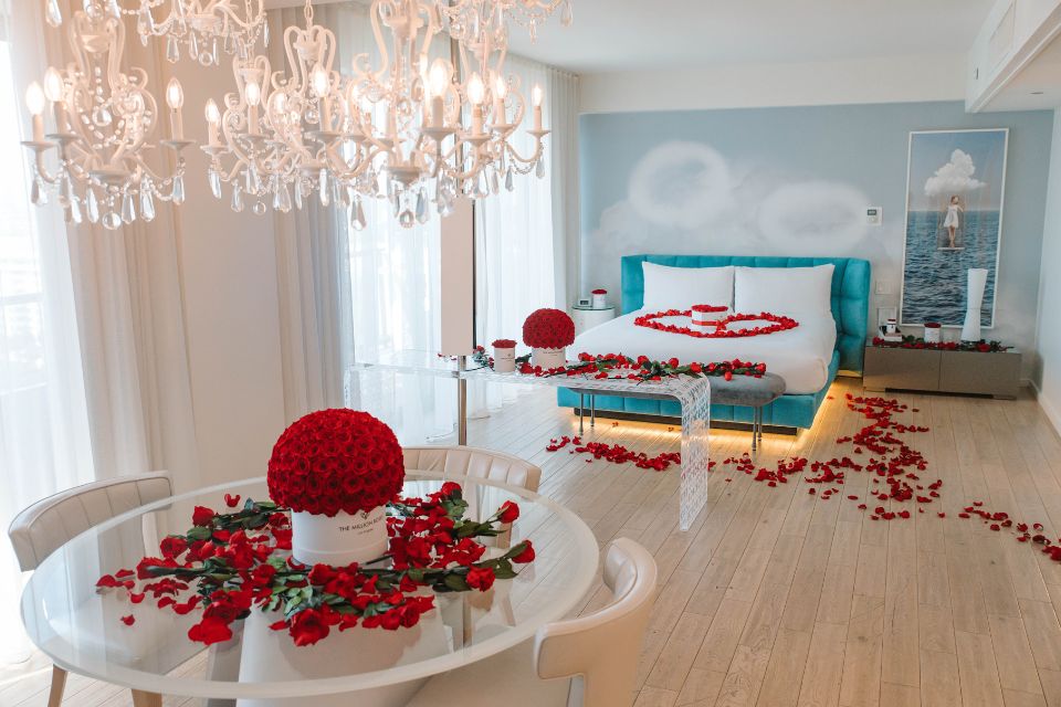 Large hotel suite with teal headboard bed with scattered rose petals all over it and the ground and a round glass table with a bouquet of roses and rose petals scattered about