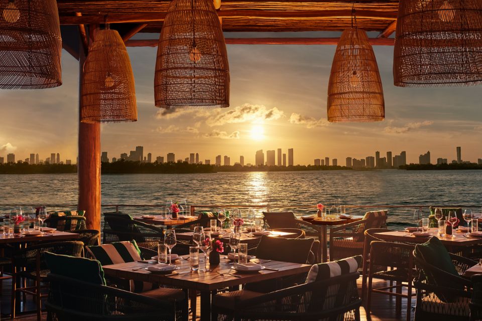 Beachfront deck with wooden tables, chairs, wicker pendant lights, and the sun setting in the distance over the ocean and city skyline