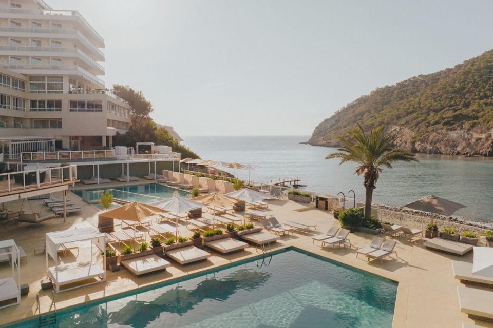 Pool deck with loung chairs and umbrellas with a white building in the background and a sea and mountain in the background