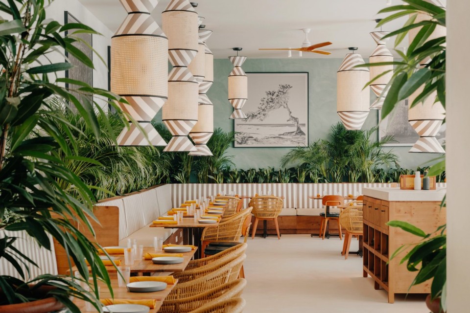 Large dining room with teal walls, many green plants, light wooden table and chairs, and striped hanging light fixtures