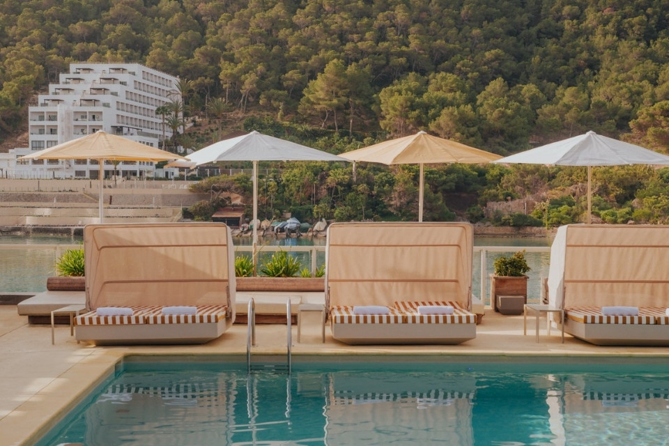 Three striped sun loungers covered with four umbrellas behind them lining a pool with a forest and white building in the background