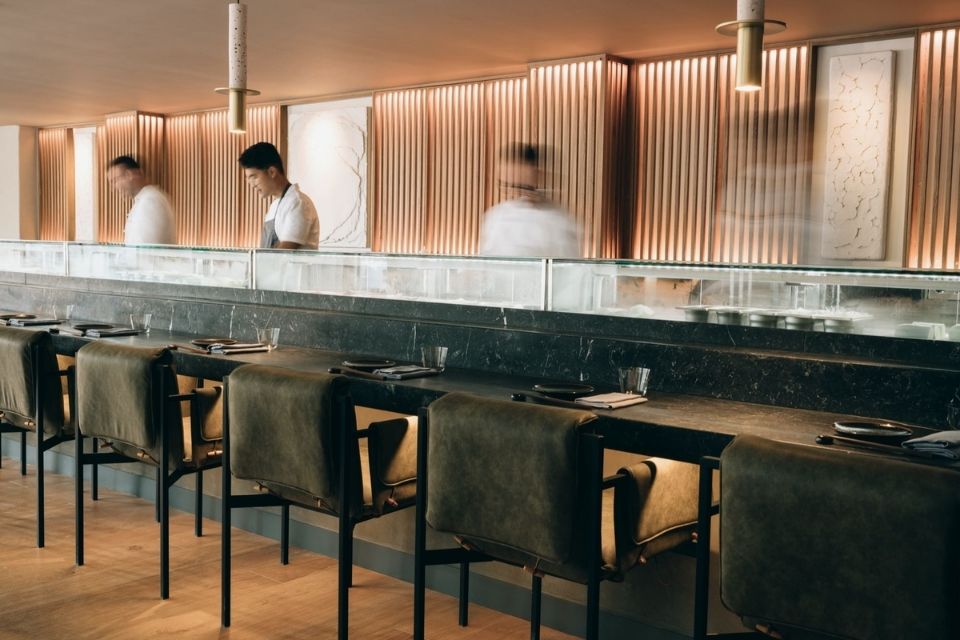 Dark green sushi counter with three sushi chefs behind it and chairs lined up in front of it