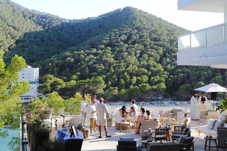 People dressed in white clothing socializing on an outdoor terrace overlooking the sea and green tree covered mountains