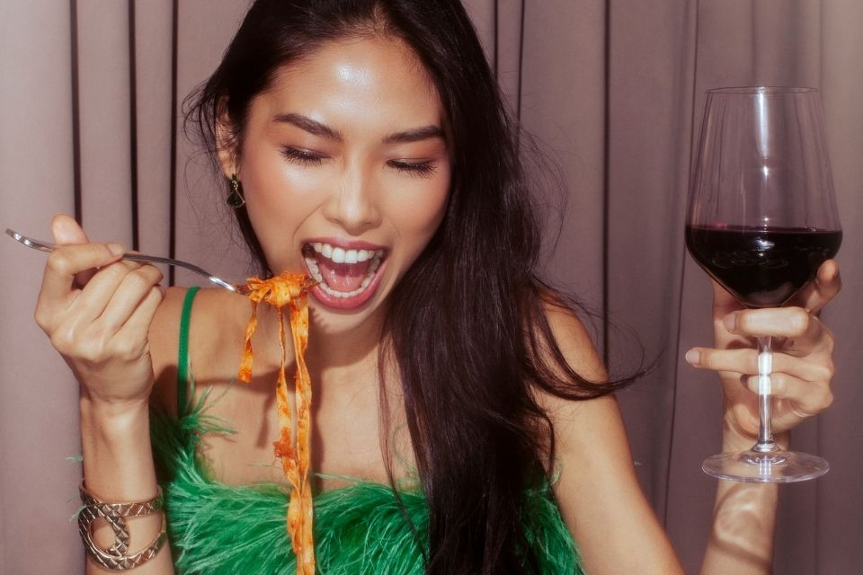 Woman with red wine in one hand and pasta on a fork in the other