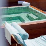 Square hot tub with turqouise tiles on a patio with a turquoise lounge chair with towels in the corner